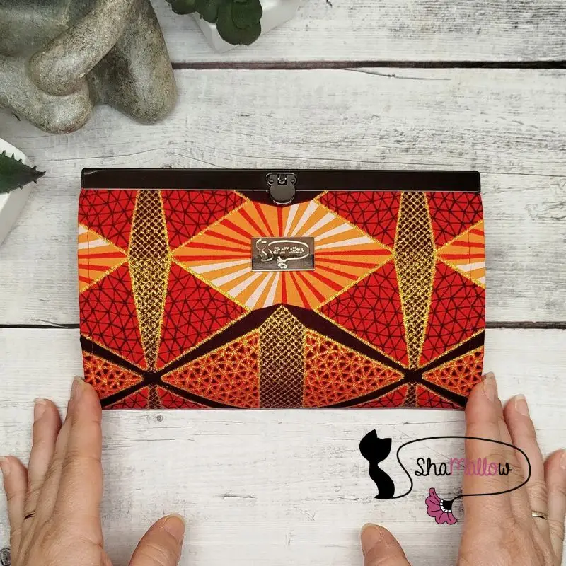 un portefeuille compagnon en tissu wax rouge orangé à paillettes dorées et motif géométrique, est tenu entre deux mains féminine sur un fond de bois blanc