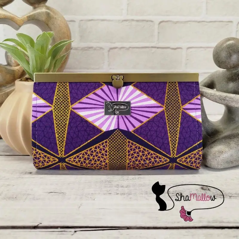 Portefeuille compagnon en tissu wax violet à paillettes dorées posé contre une petite statuette en pierre M'bigou